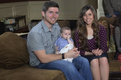 Jessa Duggar wears a purple shirt with lavender polka dots. Ben Seewald wears a grey shirt and jeans while holding their child.