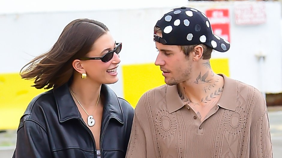 Hailey Bieber wearing a black leather jacket and sunglasses smiles at Justin Bieber who's wearing a brown shirt and backwards cap.